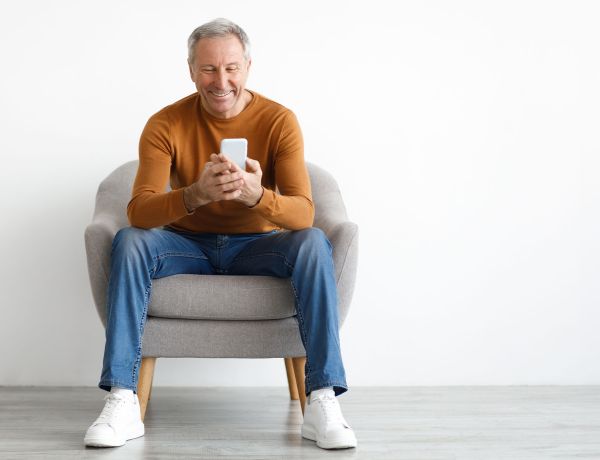 Älterer Mann mit grauen Haaren, orangefarbenem Pullover und Jeans sitzt entspannt in einem hellen Sessel und schaut lächelnd auf sein Smartphone.