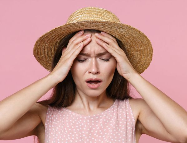 Junge Frau mit Strohhut und rosa Kleid fasst sich erschöpft an den Kopf – Symbol für Sommergrippe und Erschöpfung