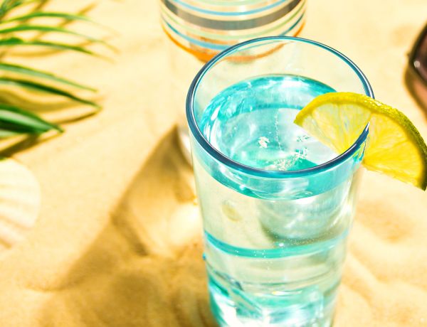 Glas Wasser mit Zitronenscheibe im Sand – erfrischendes Getränk gegen Sommerhitze