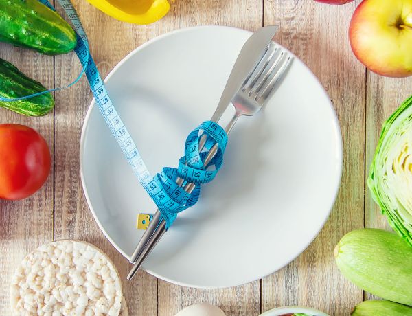 Teller mit Besteck, darauf ein Maßband, daneben frisches Gemüse und Obst als Symbol für Fastenkur.