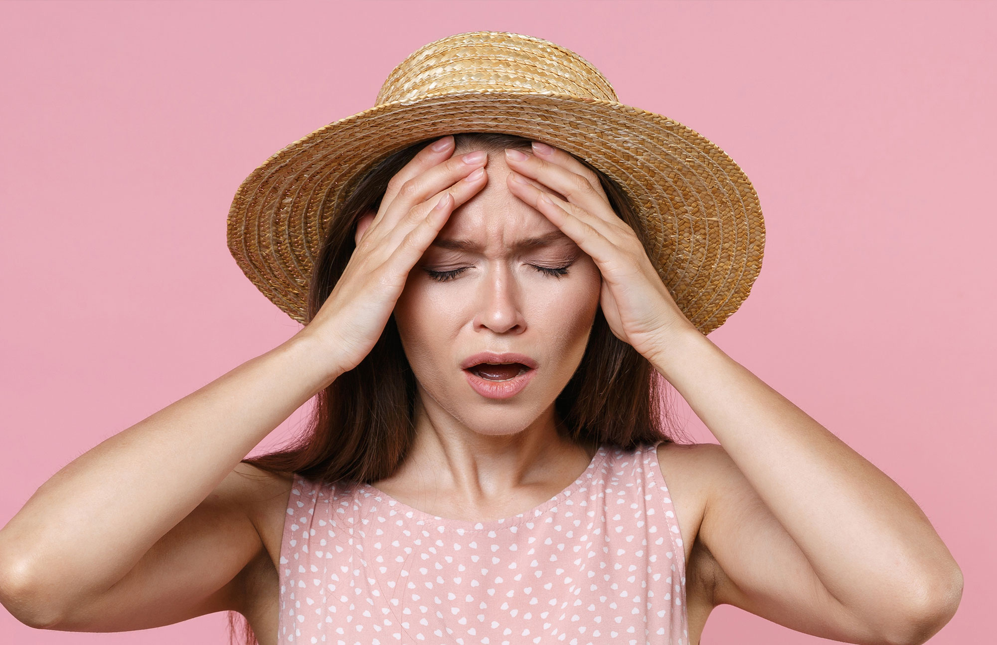 Junge Frau mit Strohhut und rosa Kleid fasst sich erschöpft an den Kopf – Symbol für Sommergrippe und Erschöpfung