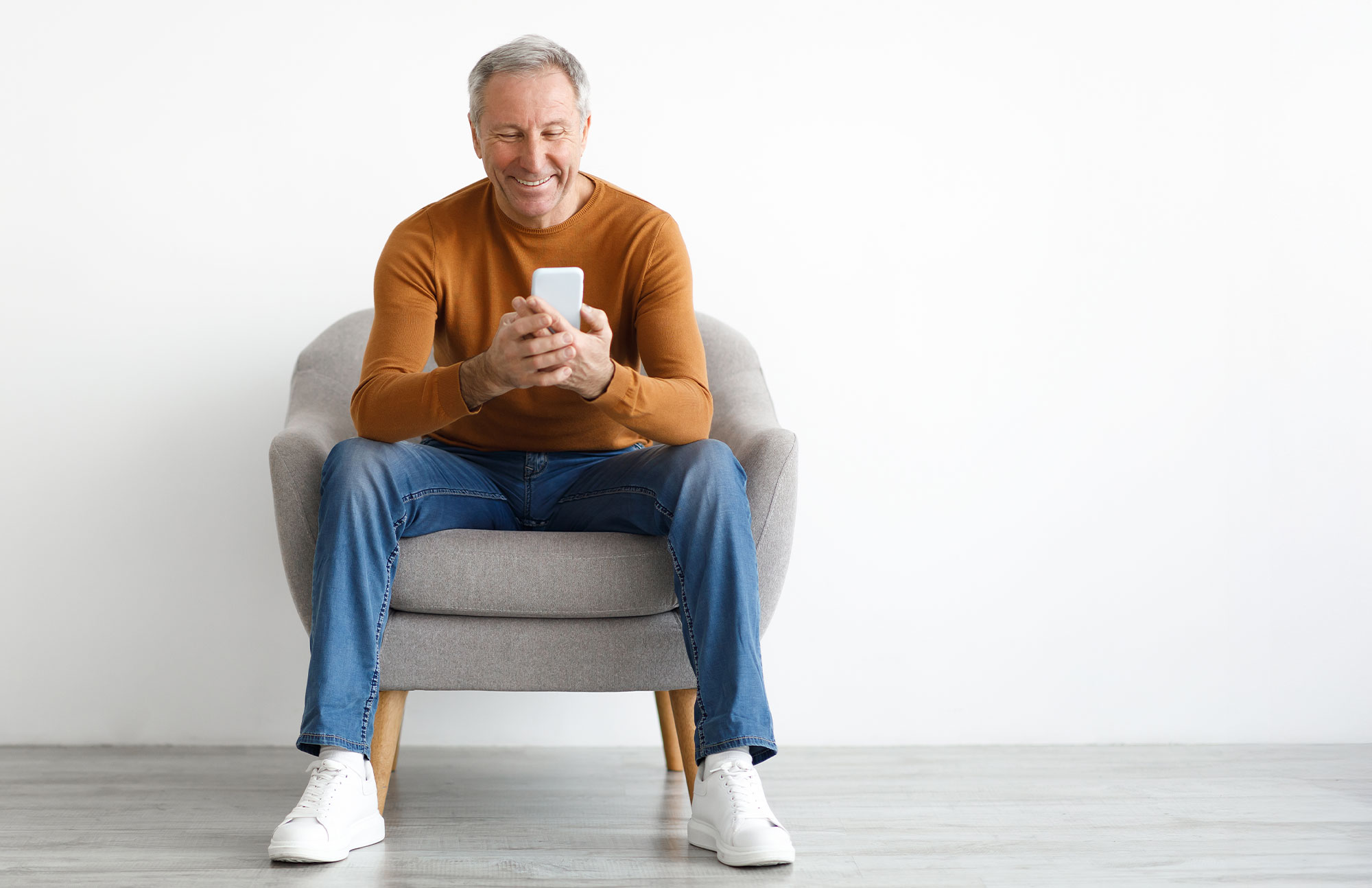 Älterer Mann mit grauen Haaren, orangefarbenem Pullover und Jeans sitzt entspannt in einem hellen Sessel und schaut lächelnd auf sein Smartphone.